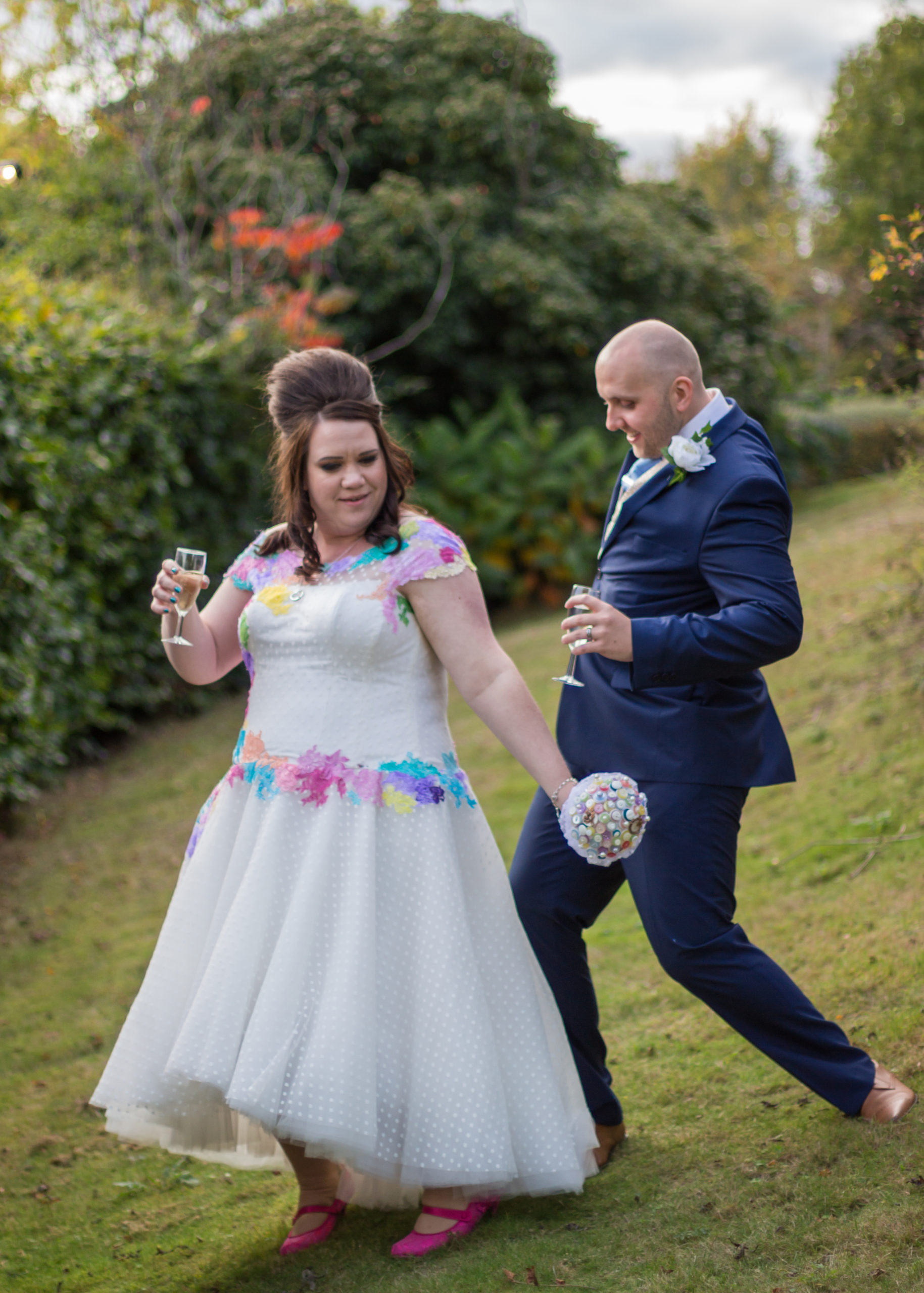 Bride holding a glass in pink shoes with groom behind her dancing
